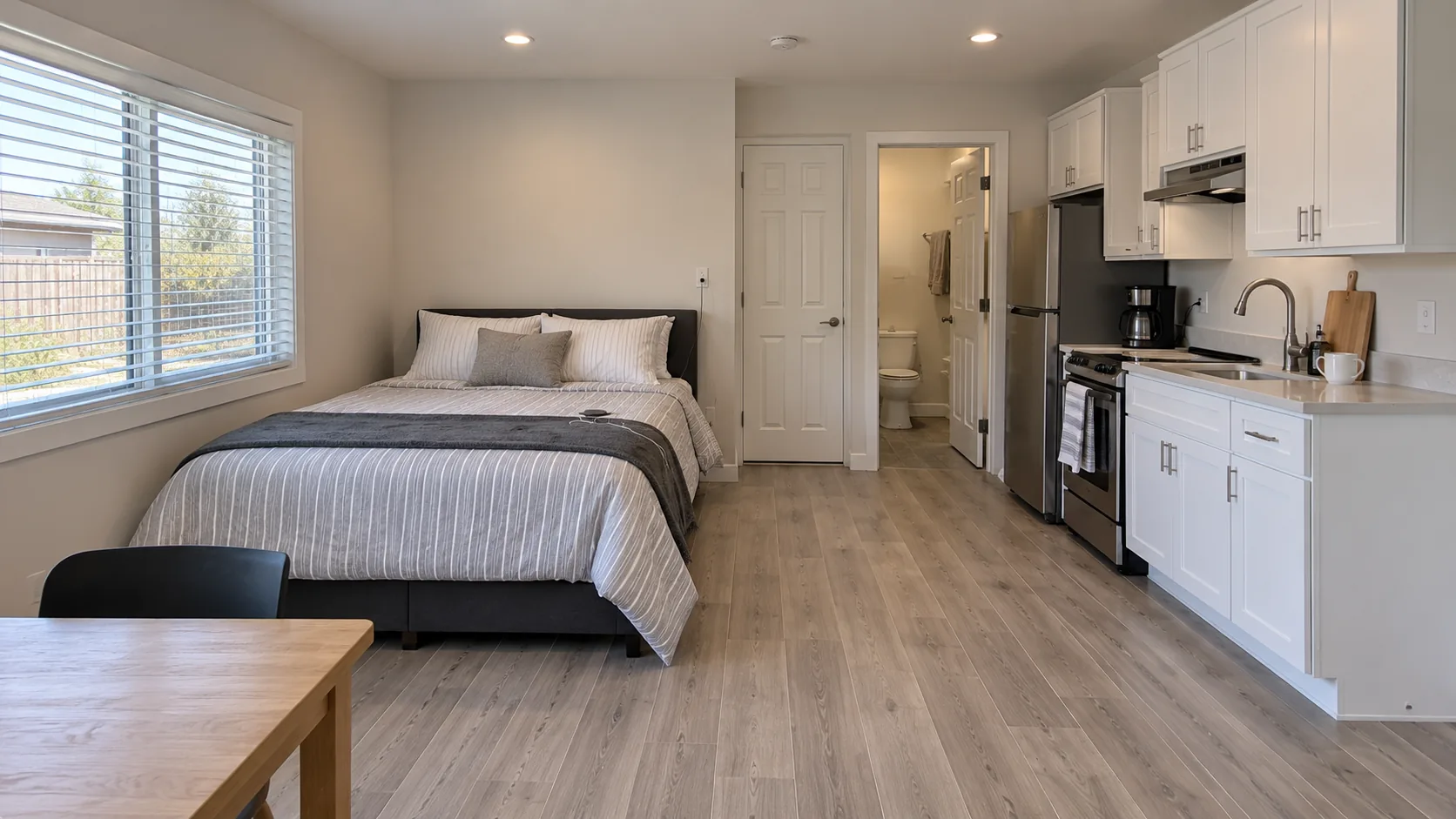 Concept image: a studio with a bed under a window and an in-line kitchen with white cabinets along one wall.