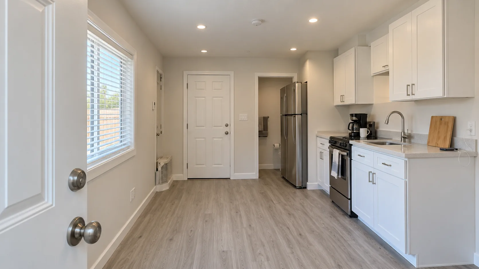 Concept image: the view from an open white door into the finished room, with a kitchen along the right wall and a bathroom door at the far end.